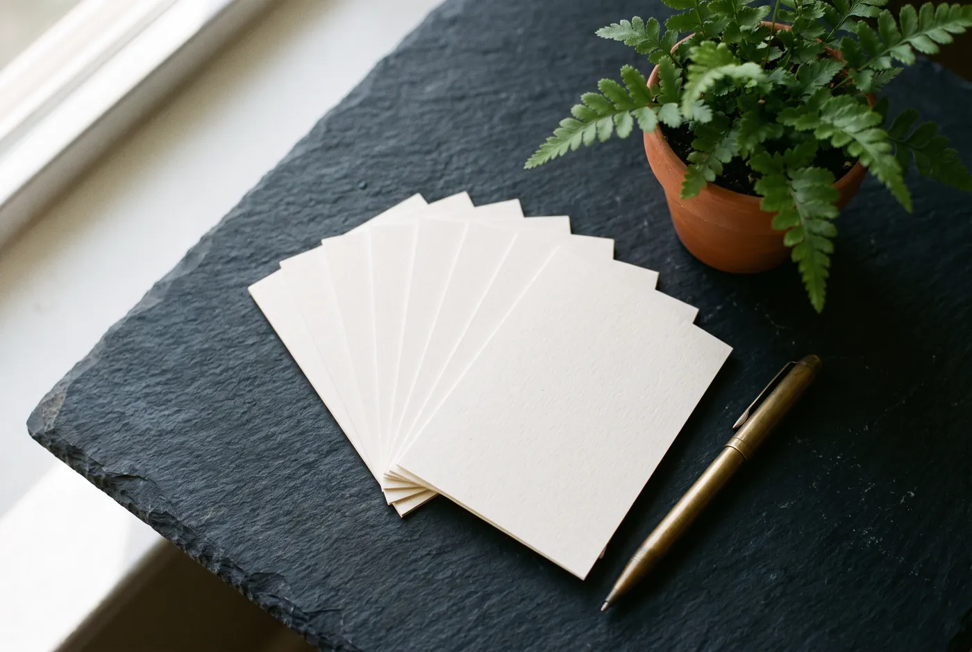 Fan of blank cream cards on a slate surface beside a fern and a brass pen