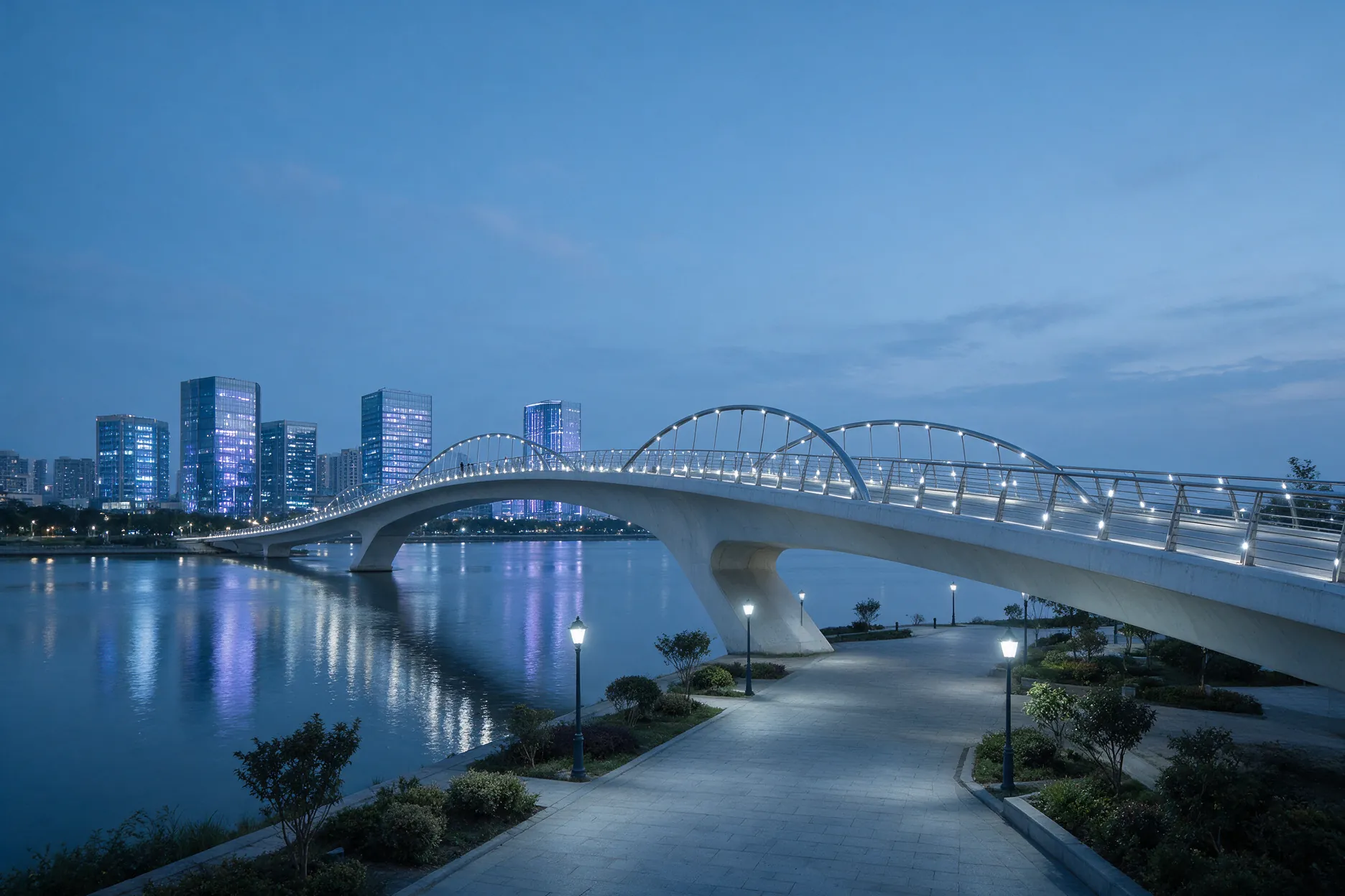 Quiet waterfront promenade and a pedestrian bridge at blue hour with lit buildings along the water